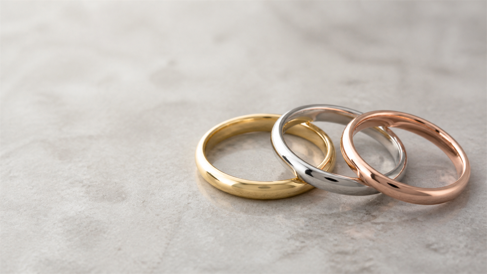 Three plain wedding bands in yellow gold, white gold or platinum, and rose gold on a grey stone surface